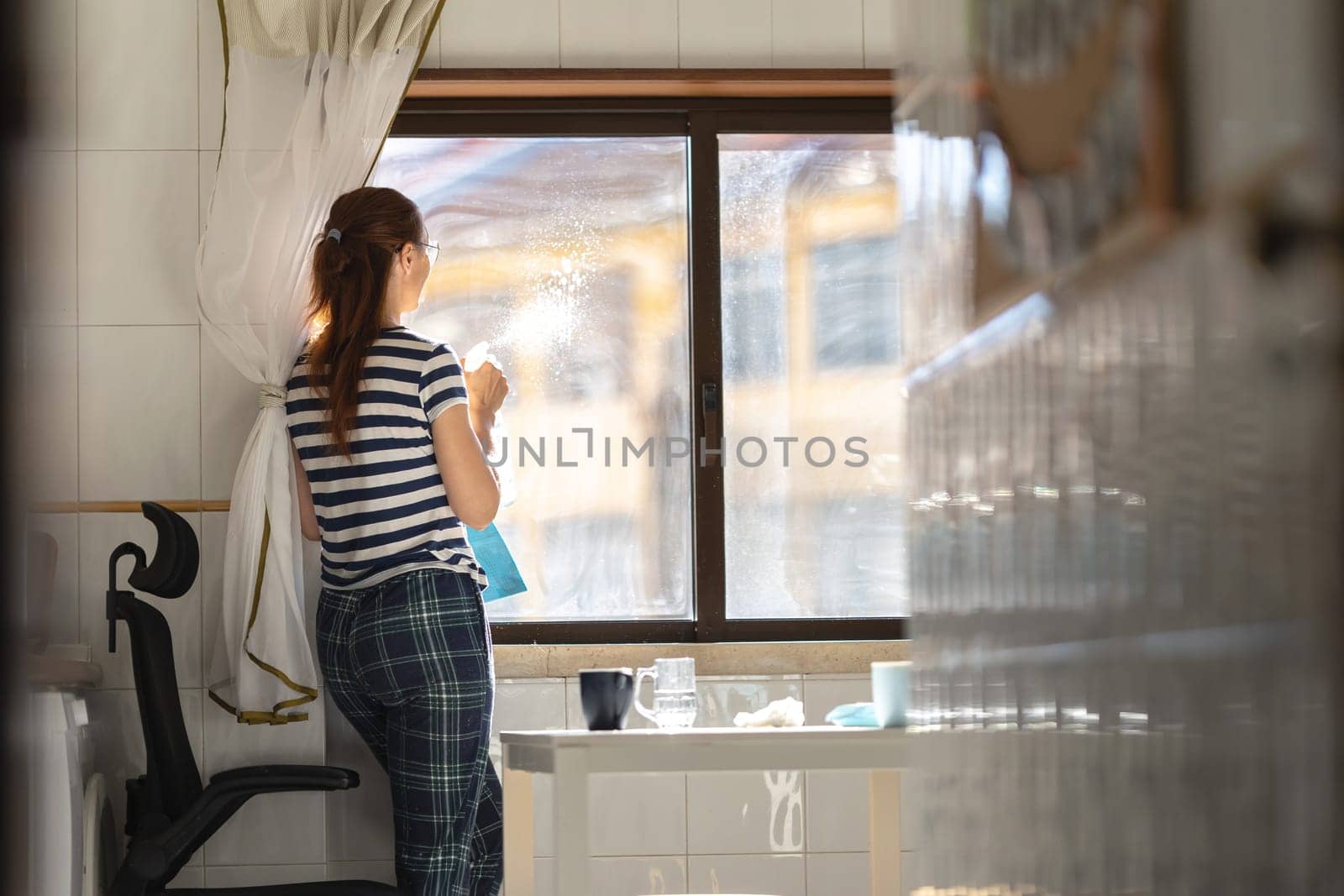 A woman housewife washing a window at the kitchen - spraying window cleaner at the glass. Mid shot