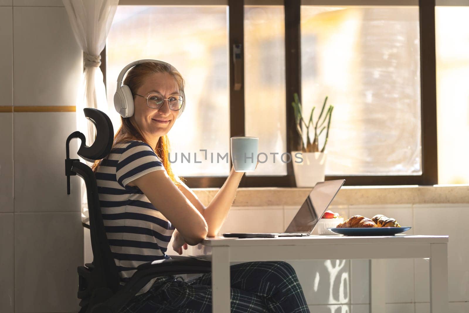 Smiling adult woman freelancer in headphones sitting by the kitchen table with a cup of coffee. Mid shot