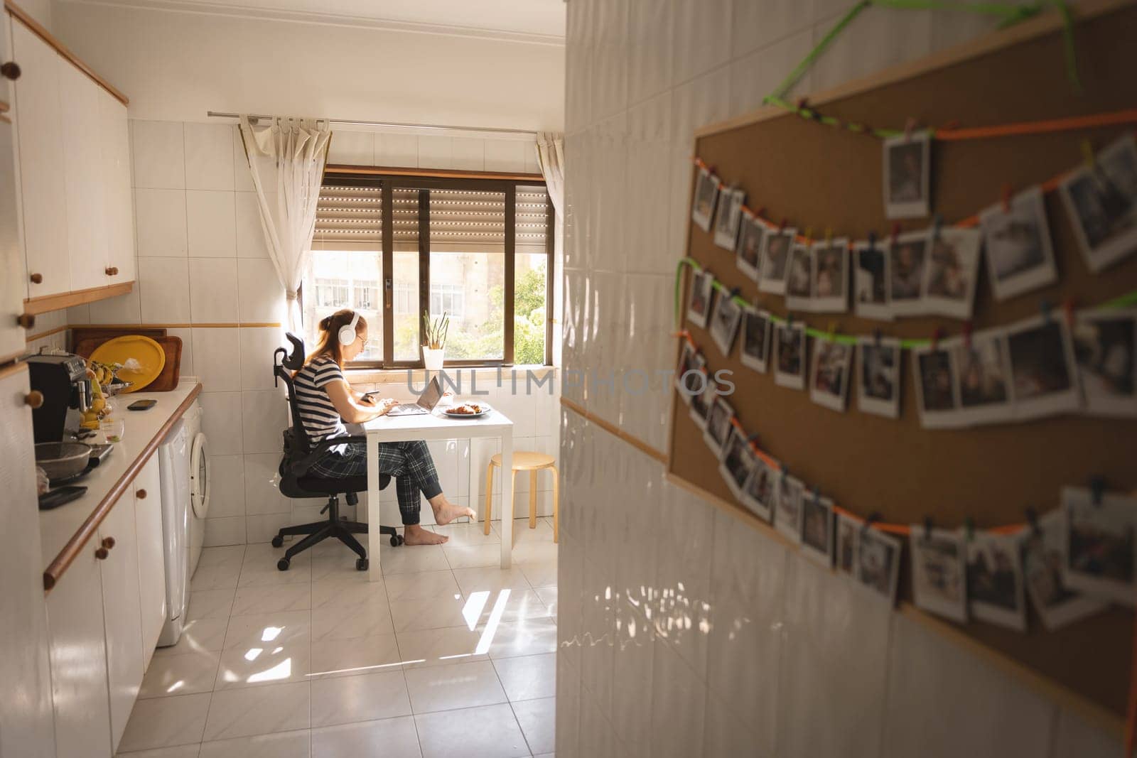 An adult business woman in headphones working at the kitchen - a board with printed photos in the foreground. Mid shot
