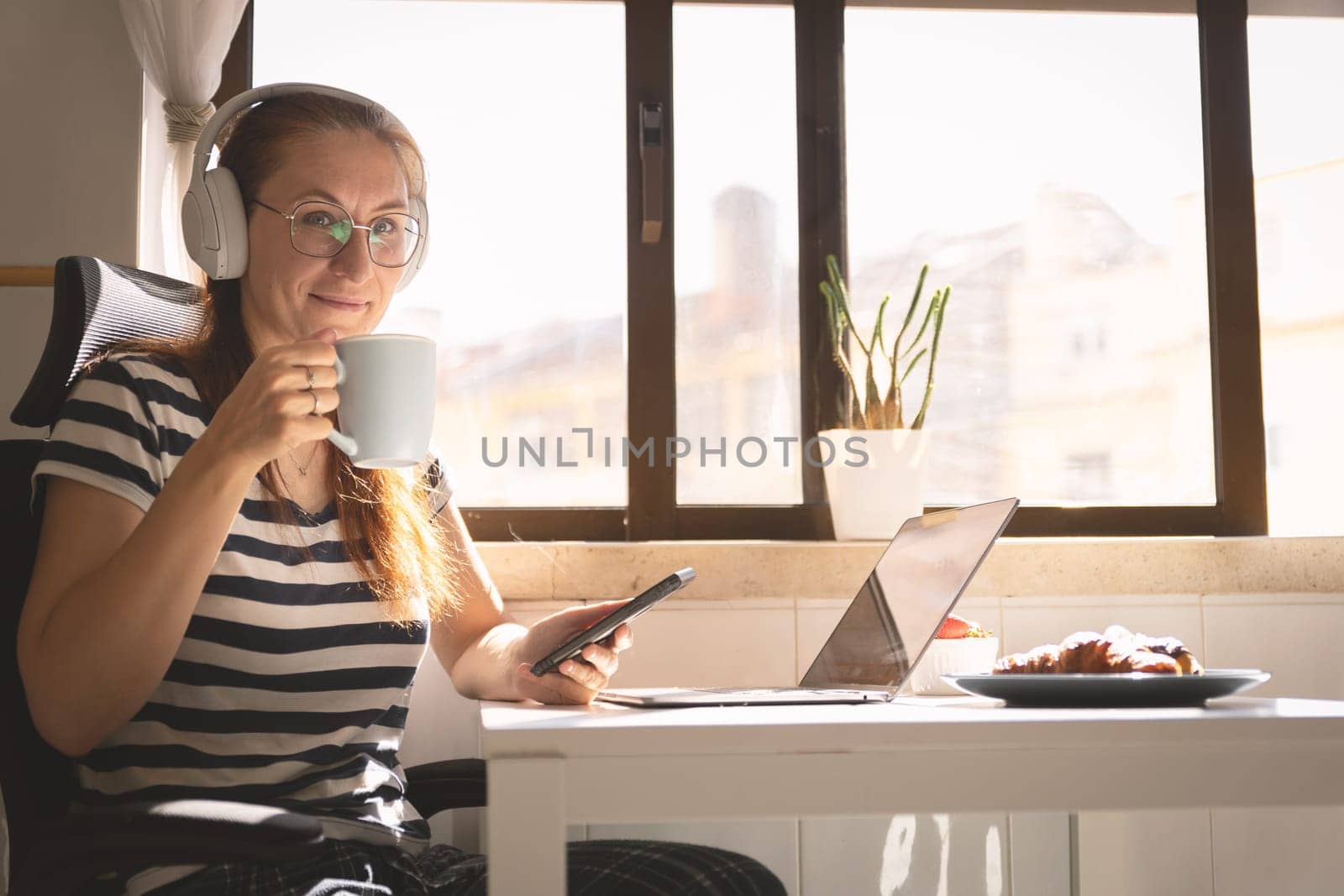 Smiling business woman in headphones sitting at the kitchen table and drinking coffee - looking in the camera. Mid shot