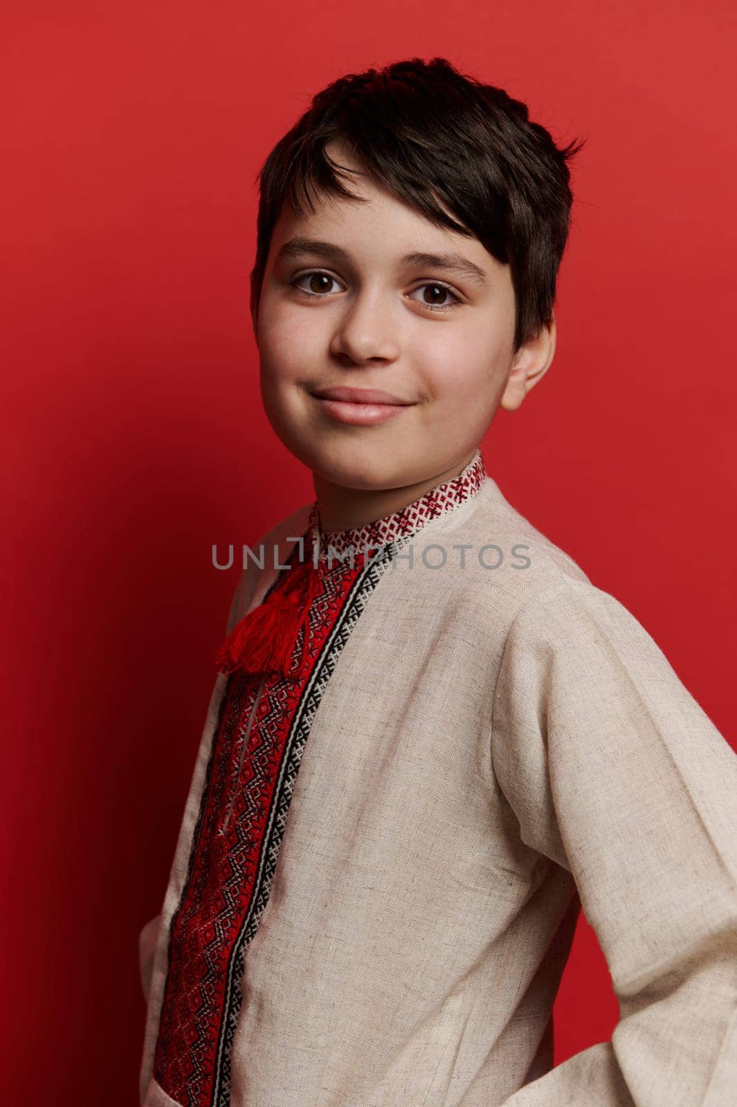 Handsome Caucasian teenage boy dressed in traditional Ukrainian embroidered linen shirt, smiling looking at camera, isolated red background. Ukrainian ethnic clothing. Fashion People Culture Folklore