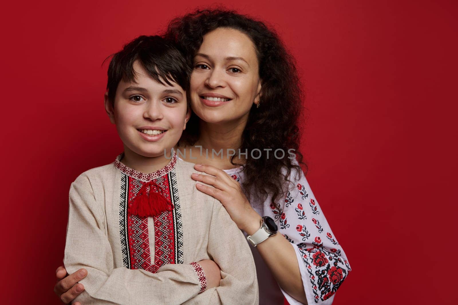 Portrait of happy mother and son wearing Ukrainian ethnic embroidered shirts, looking at camera, isolated red background by artgf