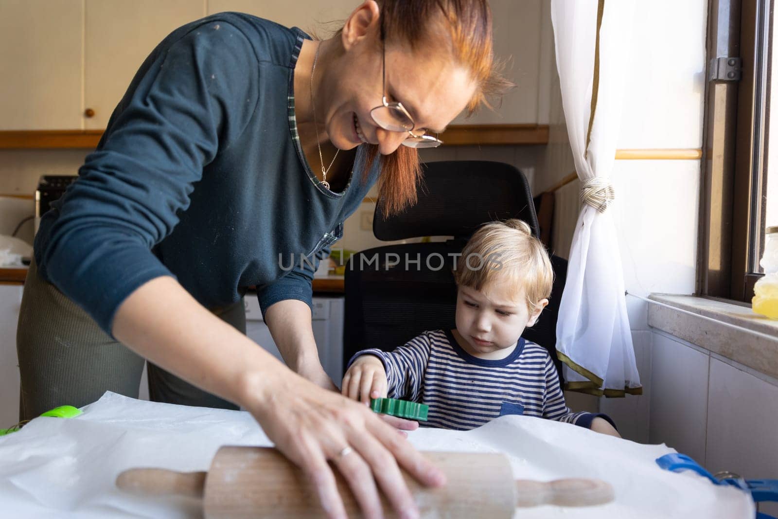 A smiling woman rolling out the dough and her little son looking at her hands. Mid shot