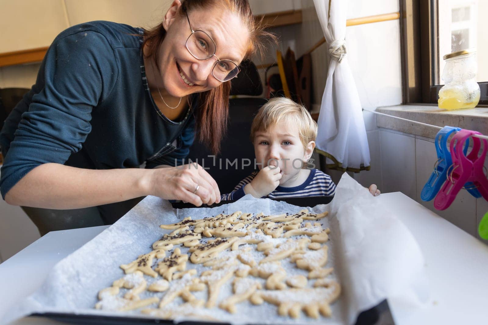 Smiling woman with her little son make cookies in the shape of dinosaurs - looking in the camera. Mid shot