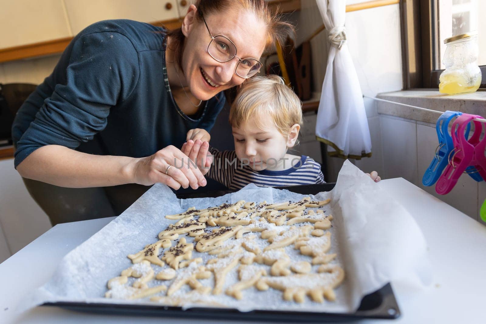 Smiling woman with her little son make cookies on the kitchen in the shape of dinosaurs - looking in the camera. Mid shot