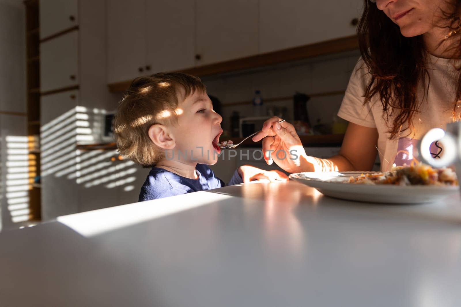 Mom feeds her little boy with a spoon in the kitchen. Mid shot