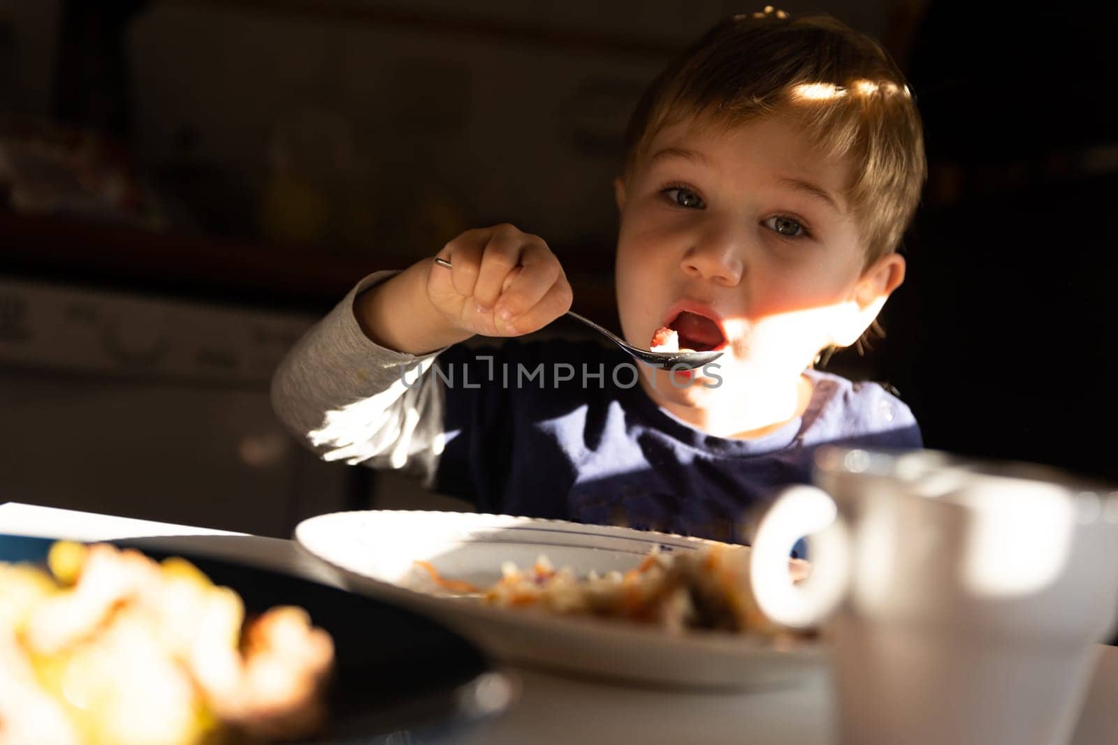 A little boy eats breakfast at the table - eats with a spoon and looks into the camera. Mid shot