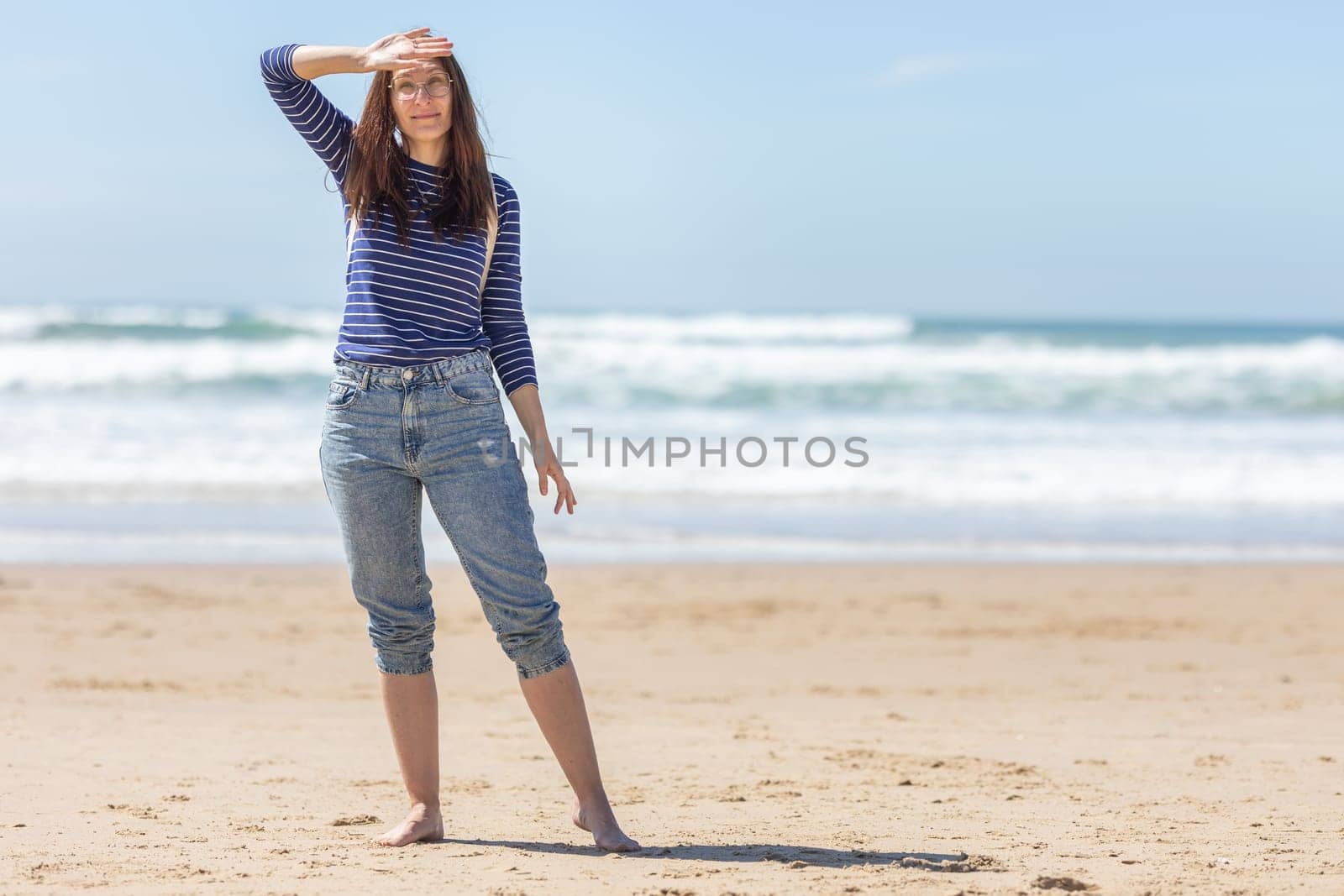 An adult smiling woman stand on sea beach and covering her face from the sunlight. Mid shot