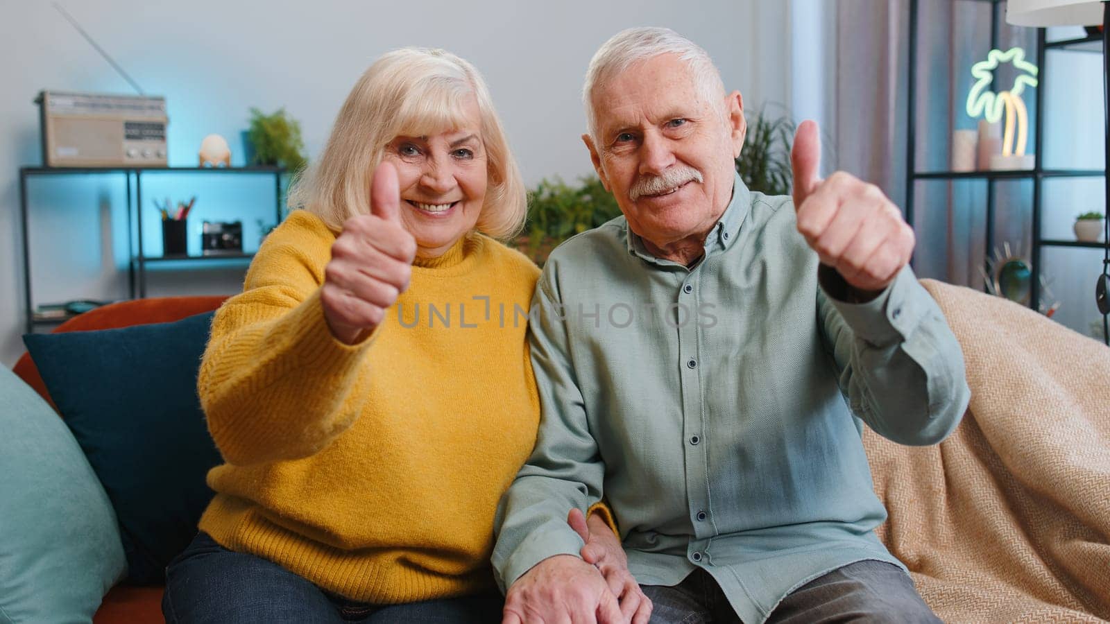 Like. Happy senior family grandparents man woman showing thumbs up, like sign positive something good, positive feedback at home. Elderly grandmother, grandfather couple sitting on sofa in living room
