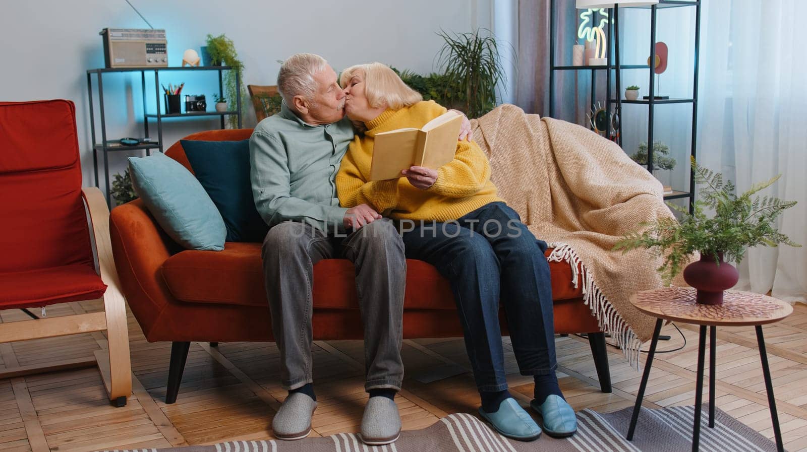 Senior family grandparents man woman reading interesting book, talking and laughing at home. Elderly grandmother, grandfather couple enjoying leisure hobbies together sitting on sofa in living room
