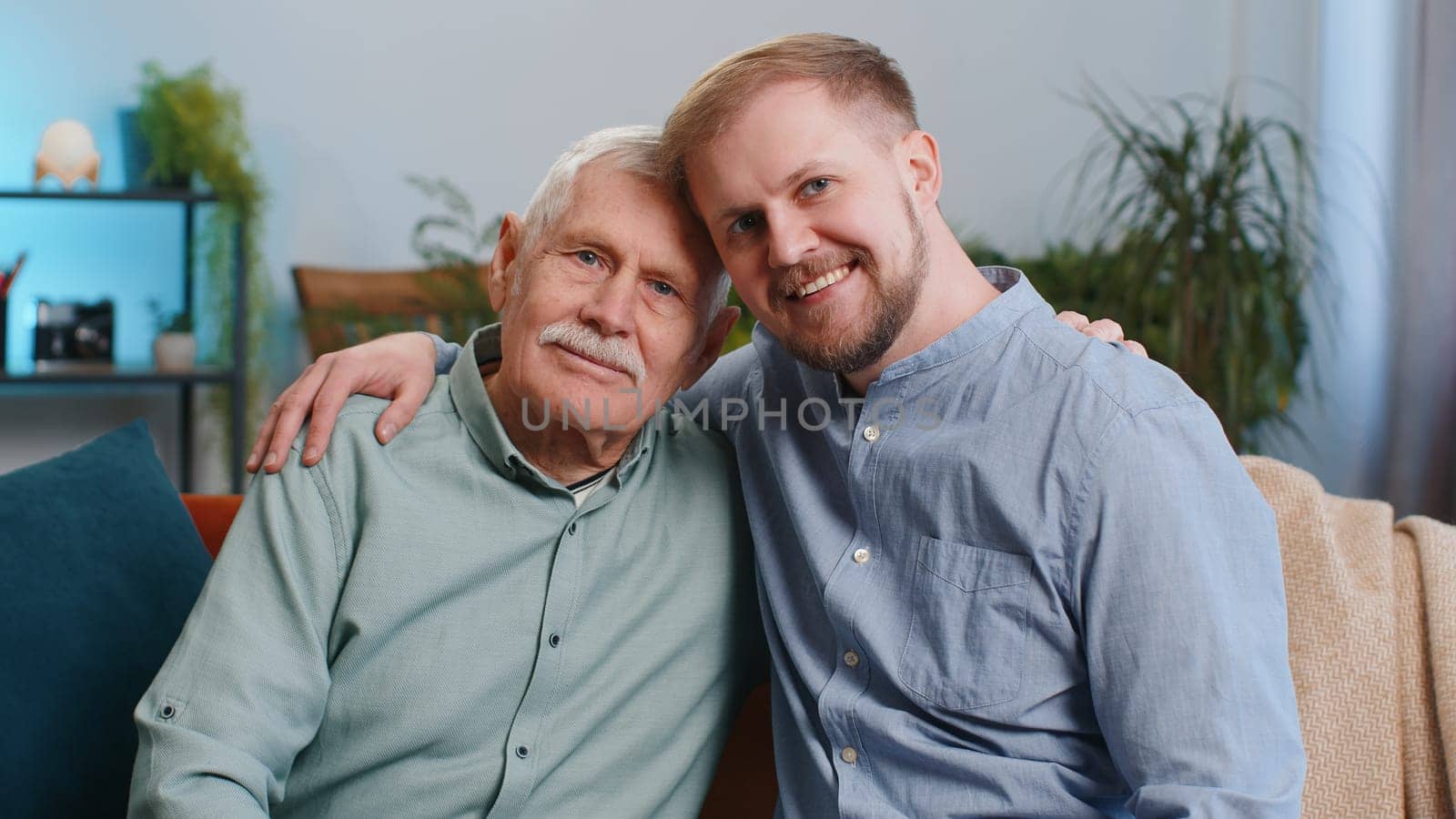 Portrait of senior grandfather with young adult man grandson smiling happy embracing hugging. Elderly father with son sitting on couch at home room. Male generations family. Bonding, good relationship