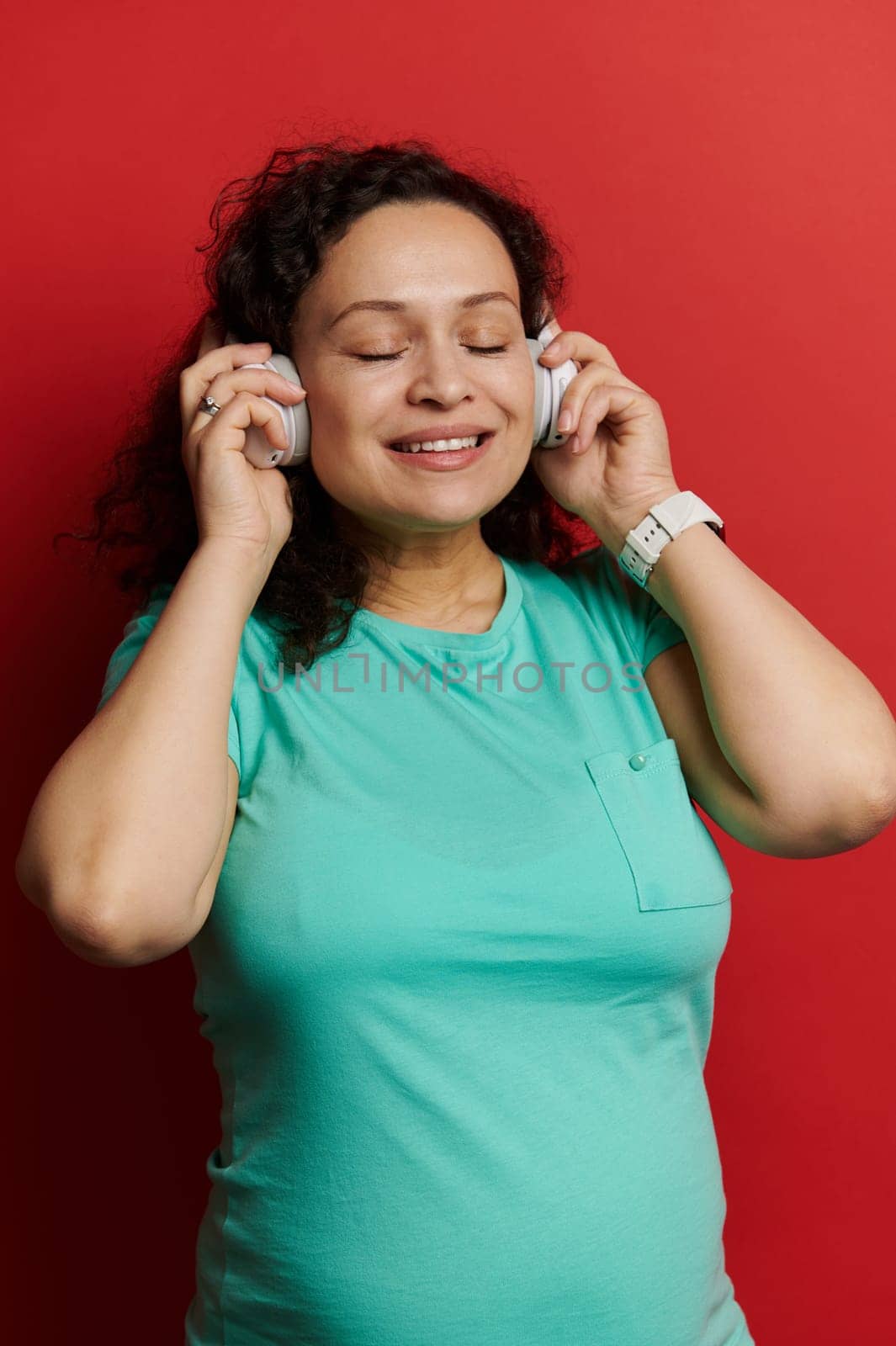 Close-up of happy pregnant positive woman with big belly, listening to music on headphones, isolated over red background by artgf