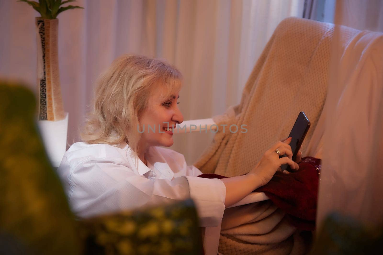 Adult mature woman of 40 or 60 years old with cell phone smartphone in casual dress white shirt and blue jeans in calm cozy evening atmosphere room. Interior with curtains and soft warm lamp