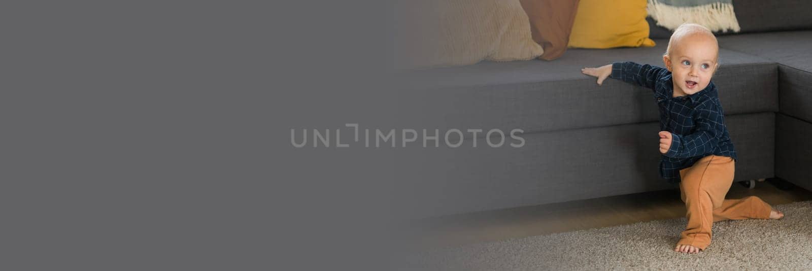 Banner Toddler boy laughing having fun standing near sofa in living room at home copy space. Adorable baby making first steps alone. Happy childhood and child care concept by Satura86