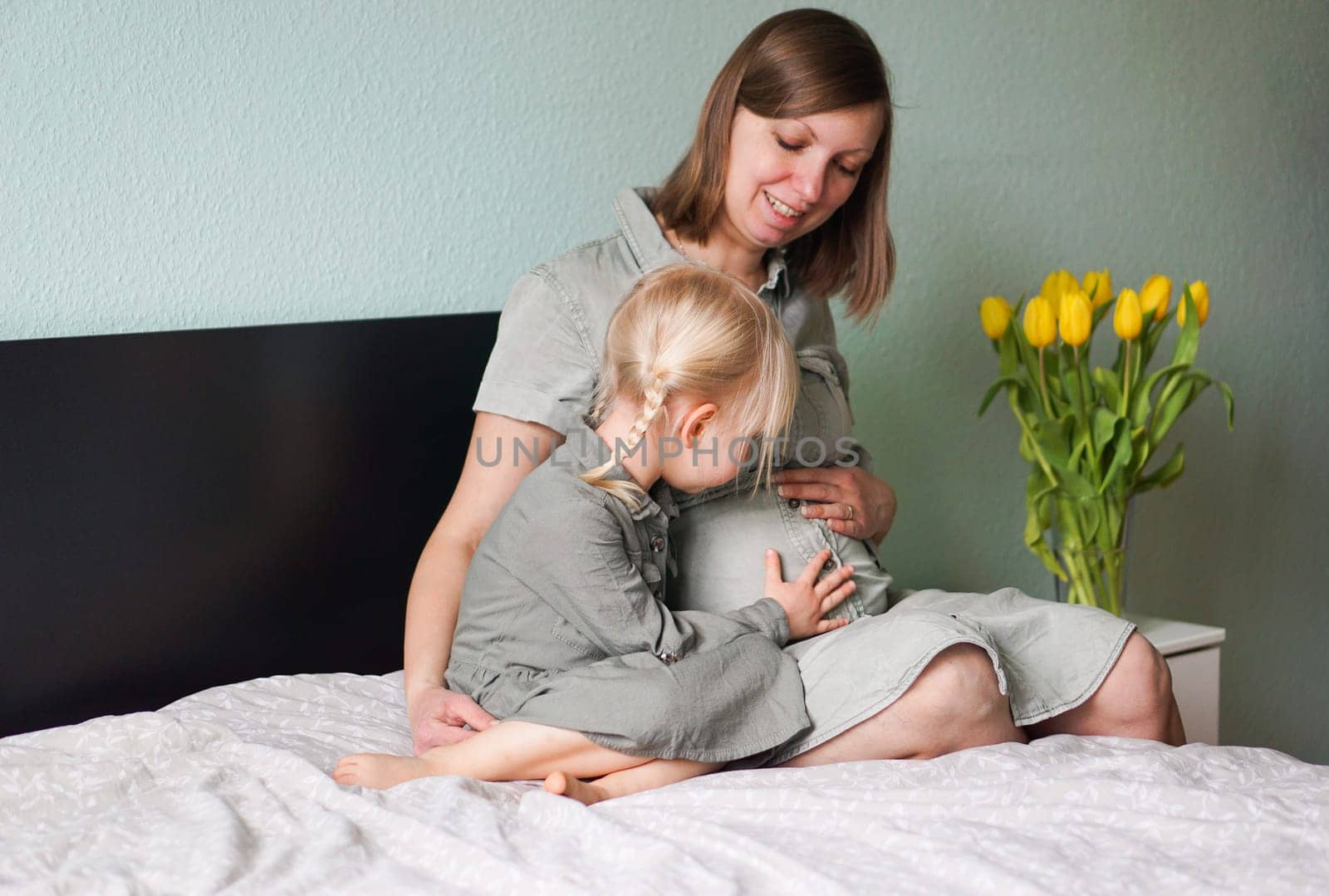 Beautiful smiling pregnant woman with her little daughter are talking and spending time together at home. Lovely child touching mothers pregnant belly. Expecting mother sitting with her cute girl.