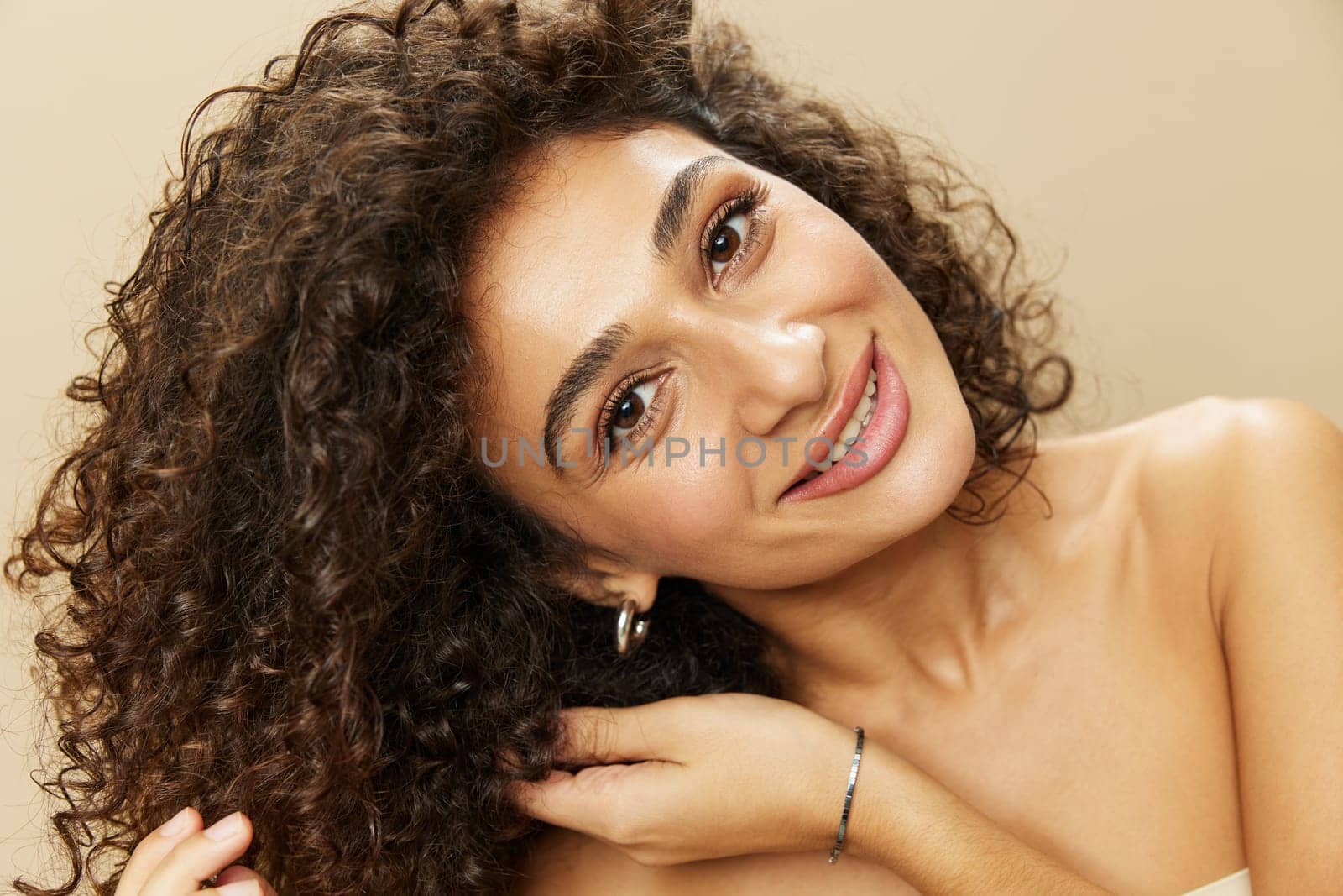 Woman applies cream and balm to her curly hair, the concept of protection and care, a healthy look, a smile with teeth on a beige background.