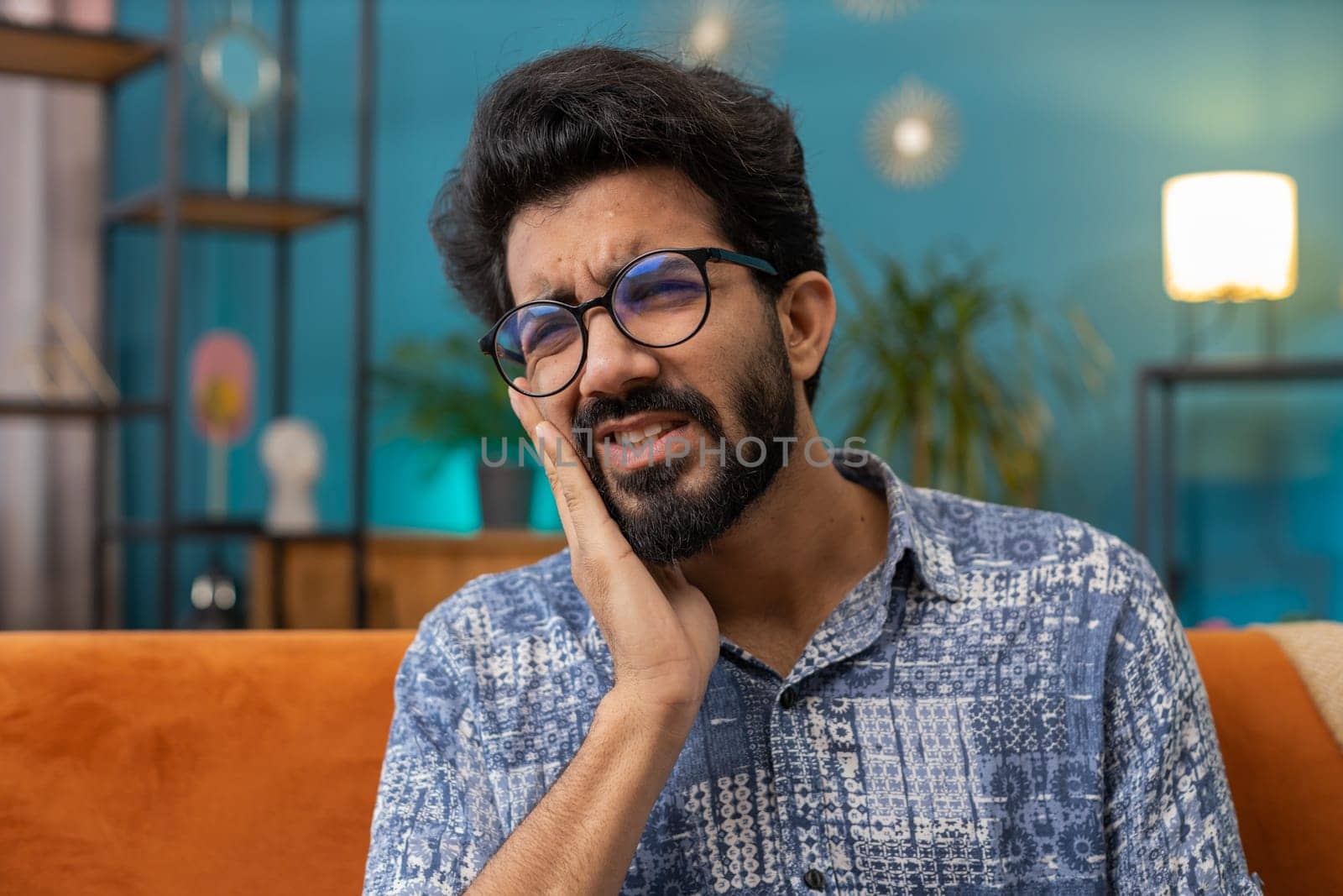 Dental problems. Young indian man touching cheek, closing eyes with expression of terrible suffer from painful toothache, sensitive teeth, cavities. Hindu guy at home apartment living room on sofa