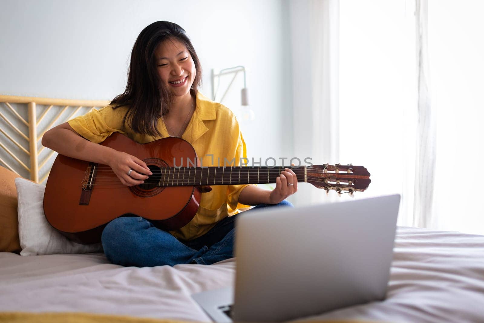 Happy teenage asian girl learning to play guitar with online teacher using laptop in bedroom sitting on bed. Copy space. by Hoverstock