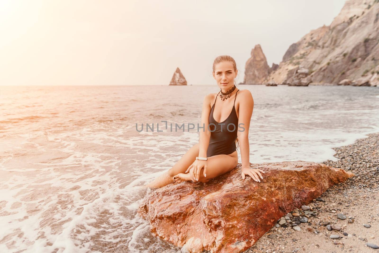Woman travel portrait. Happy woman with long hair looking at camera and smiling. Close up portrait cute woman in black bikini posing on a red volcanic rock on the sea beach by panophotograph