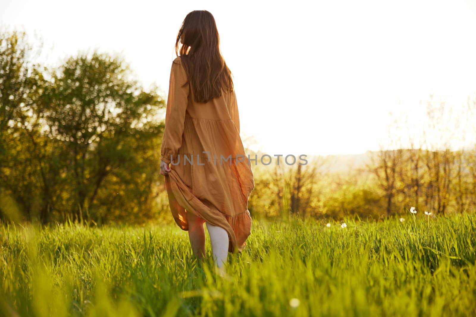 a slender woman stands in a long orange dress with her back to the camera illuminated by the sunset rays of the sun and looks towards the sky by Vichizh