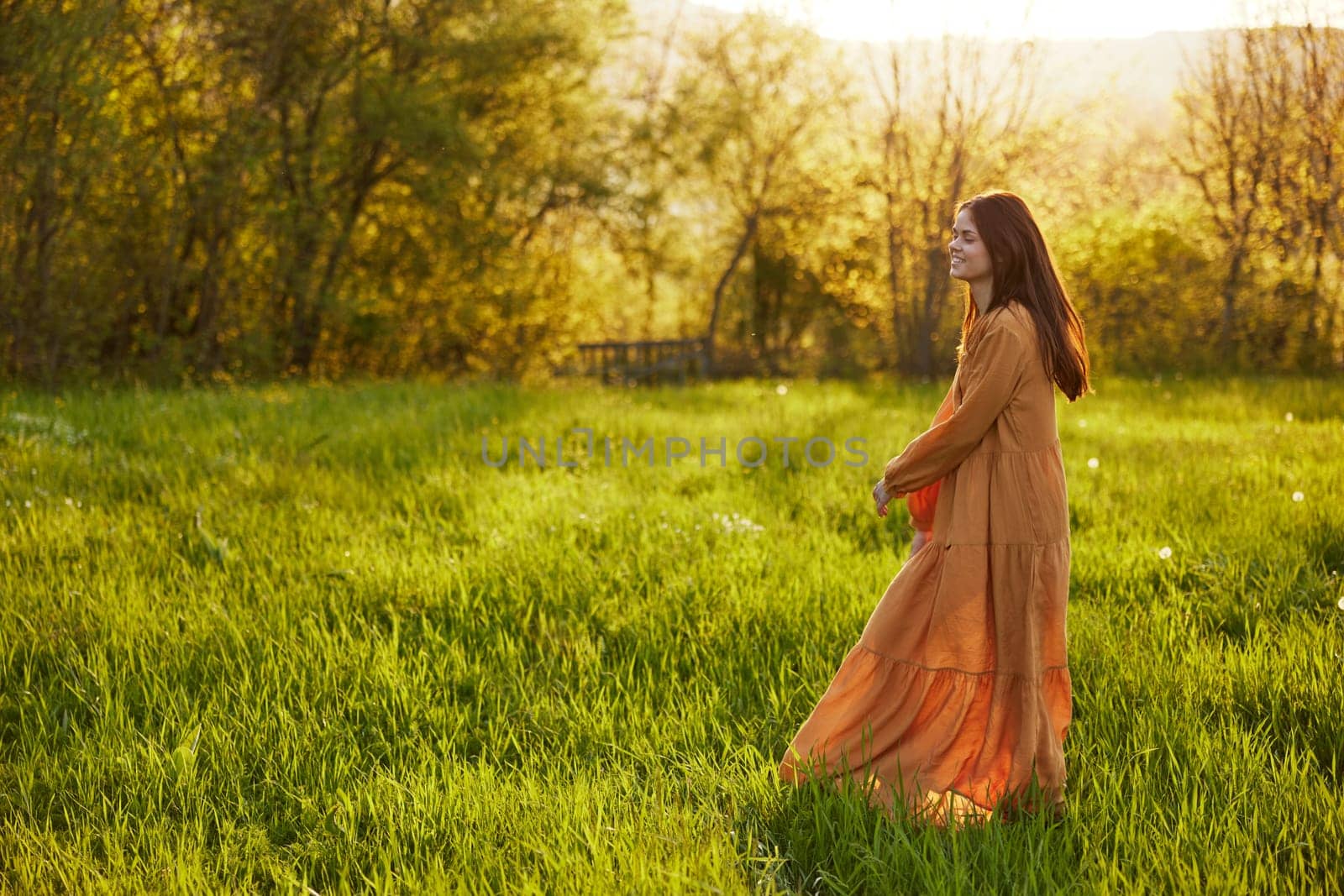 a relaxed slender woman enjoys the sunset standing in a green field with tall grass in an orange dress posing looking away by Vichizh