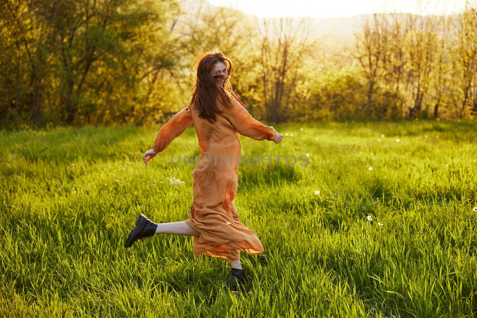 a happy woman in an orange dress illuminated by the rays of the setting sun is happily spinning in a green field, enjoying nature on a warm summer day by Vichizh