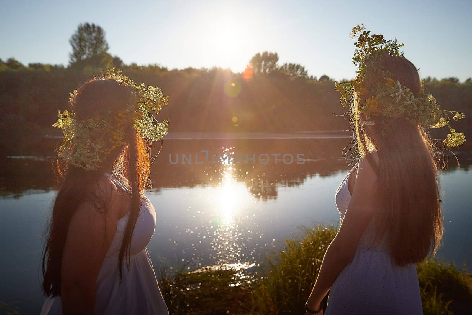 Adult mature and young brunette women in white sundresses and wreaths of flowers in summer by water of river or lake at sunset. Girls mother and daughter Celebrating pagan holiday of Ivan Kupala by keleny