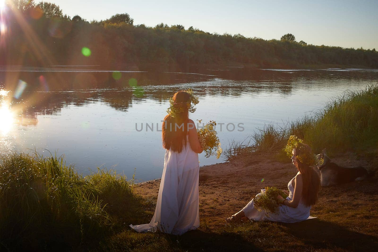 Adult mature and young brunette women in white sundresses and wreaths of flowers in summer by water of river or lake at sunset. Girls mother and daughter Celebrating pagan holiday of Ivan Kupala by keleny