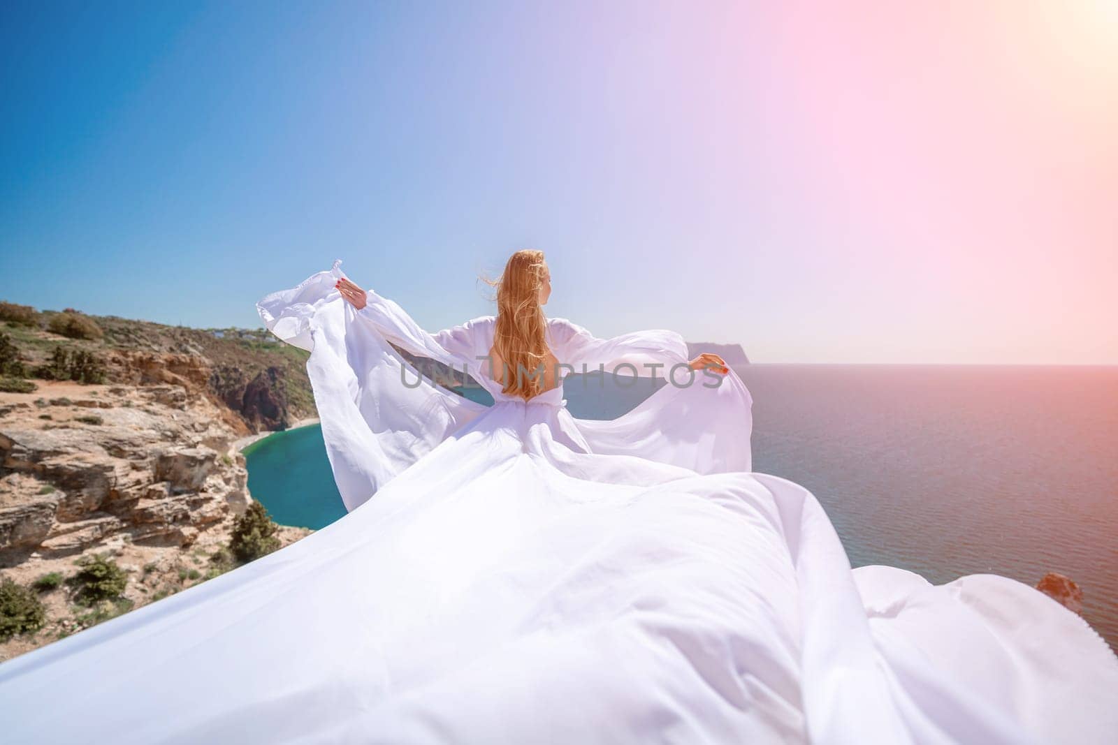 woman sea white dress. Blonde with long hair on a sunny seashore in a white flowing dress, rear view, silk fabric waving in the wind. Against the backdrop of the blue sky and mountains on the seashore