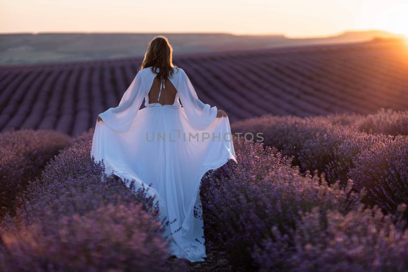 Happy woman in a white dress and straw hat strolling through a lavender field at sunrise, taking in the tranquil atmosphere