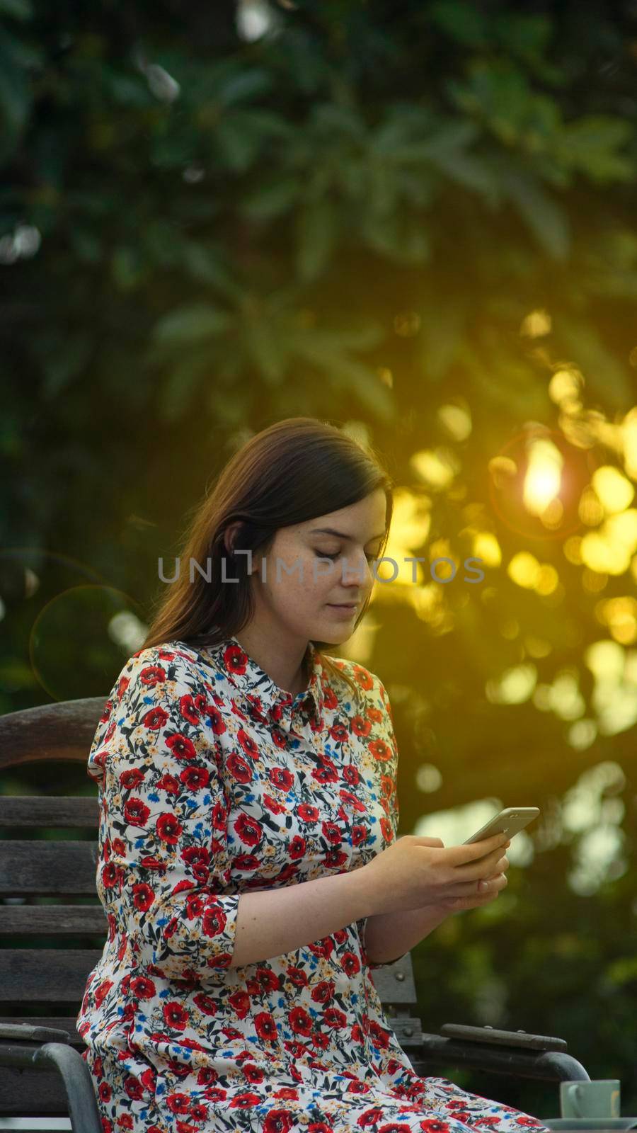 Beautiful young latin woman in white floral design dress reading and typing on her smartphone in the garden at sunset with the sun's rays passing through the trees in the background