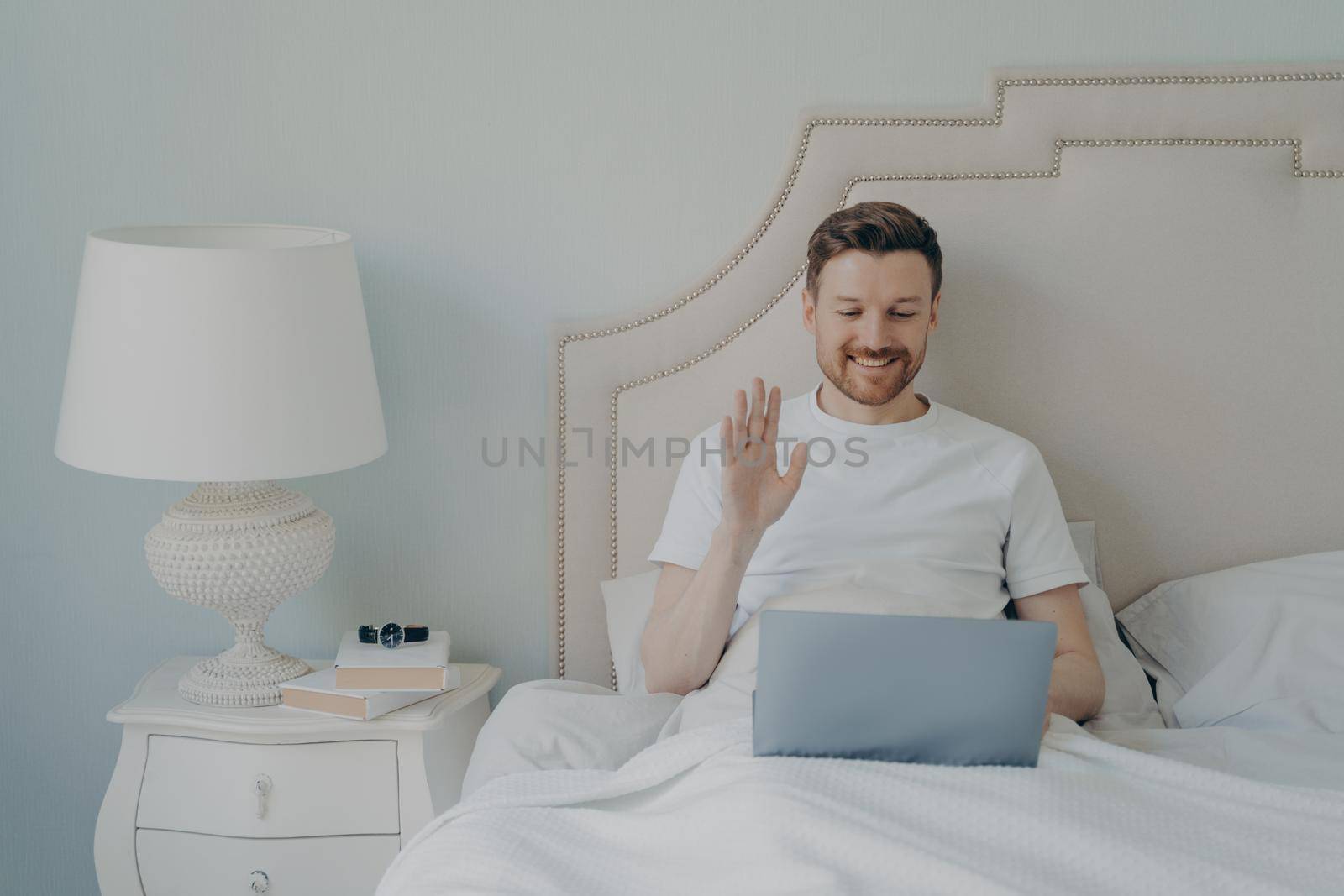 Relaxed young man with stubble using laptop laptop while sitting in bed by vkstock