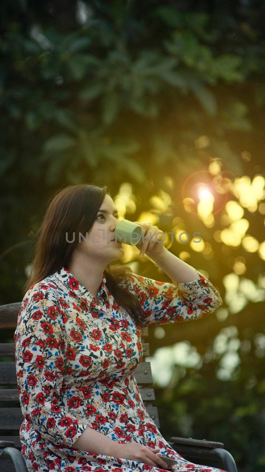 Beautiful latin young woman in white floral design dress drinking a cup of coffee in the garden at sunset with the sun's rays passing through the trees in the background