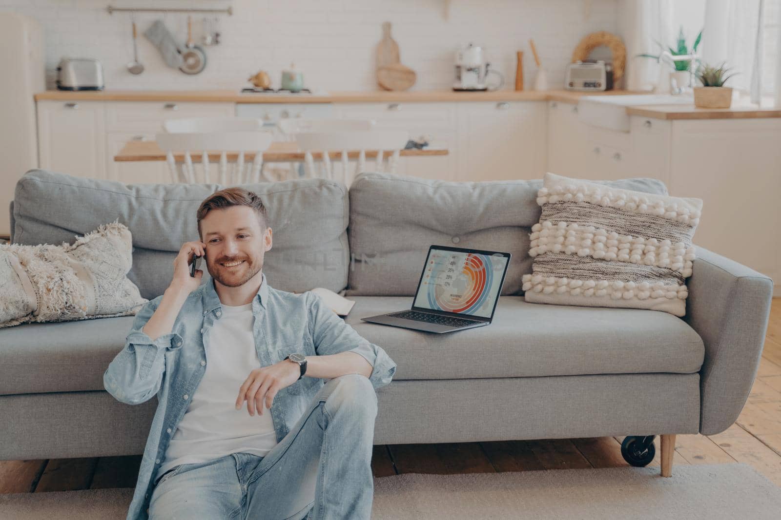 Confident male freelance worker calling his employer to tell him good news about project, making another client satisfied, laptop with infographics, sitting on floor while resting against couch