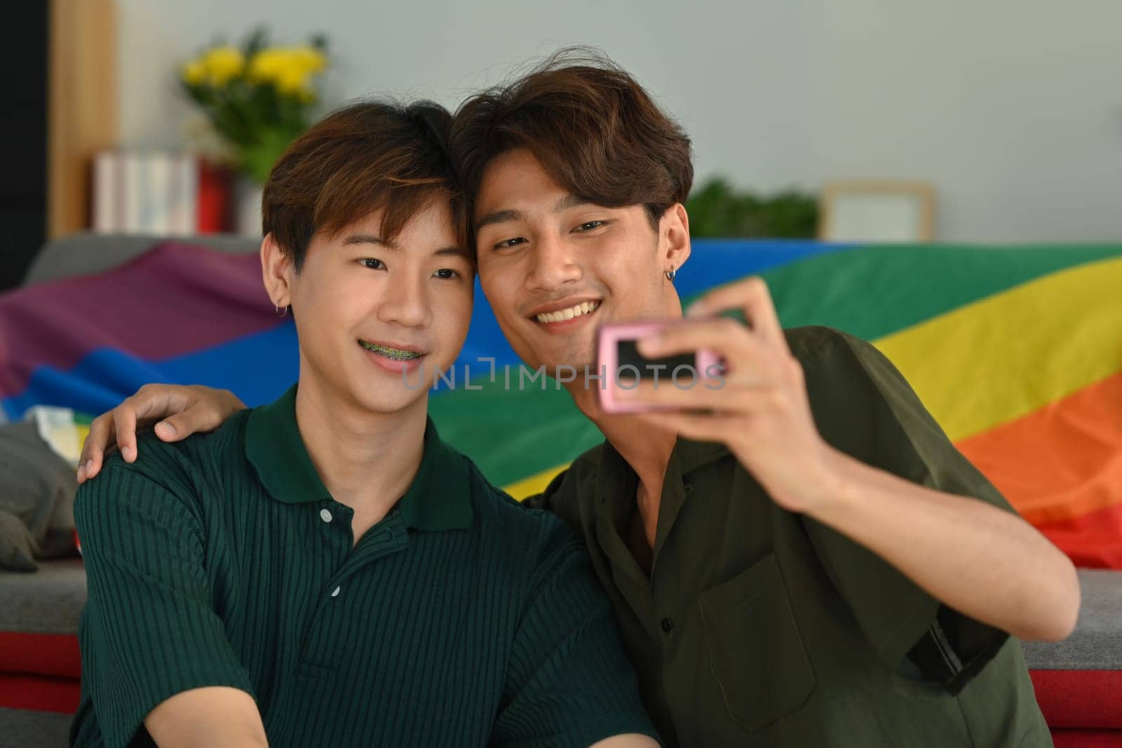 Joyful gay couple taking a selfie with compact camera in living room rainbow flag in background. LGBT, love and human rights concept.