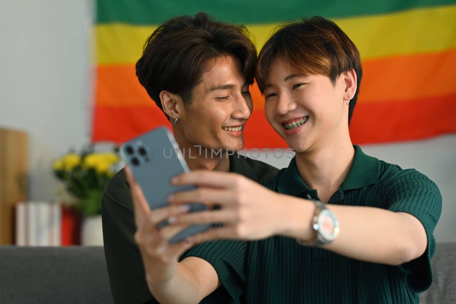 Romantic young gay couple taking a selfie with smartphone in living room rainbow flag in background. LGBT and love concept.