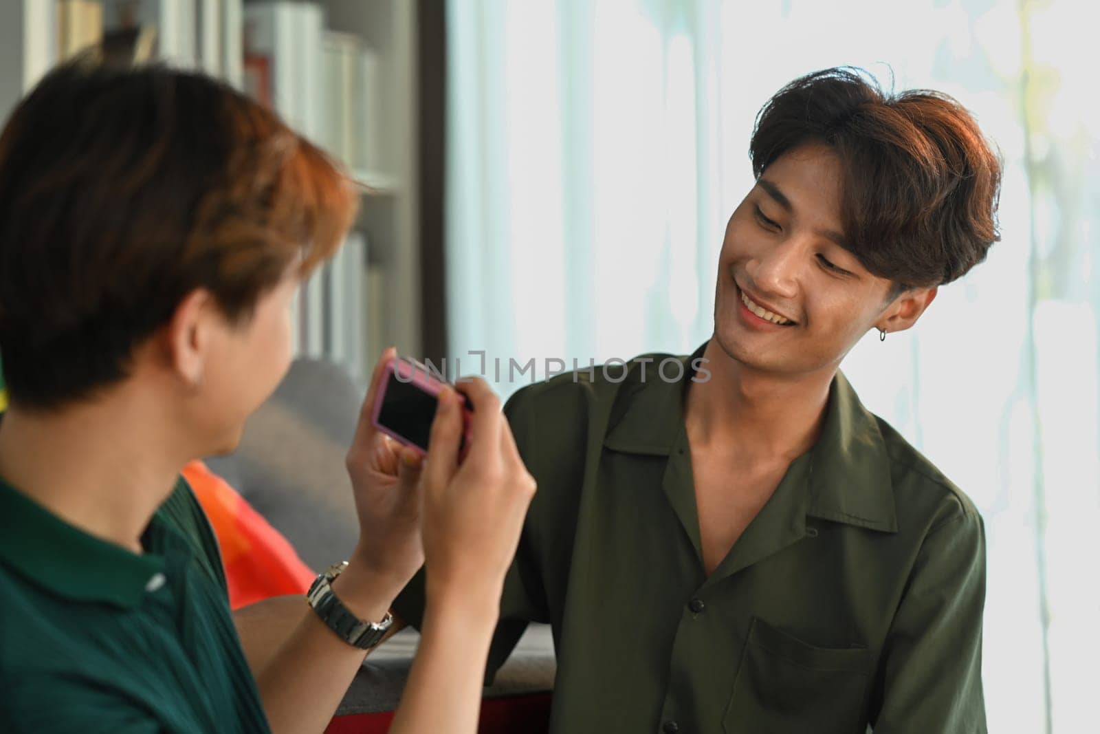 Joyful gay couple taking a selfie with compact camera in living room rainbow flag in background. LGBT, love and human rights concept.