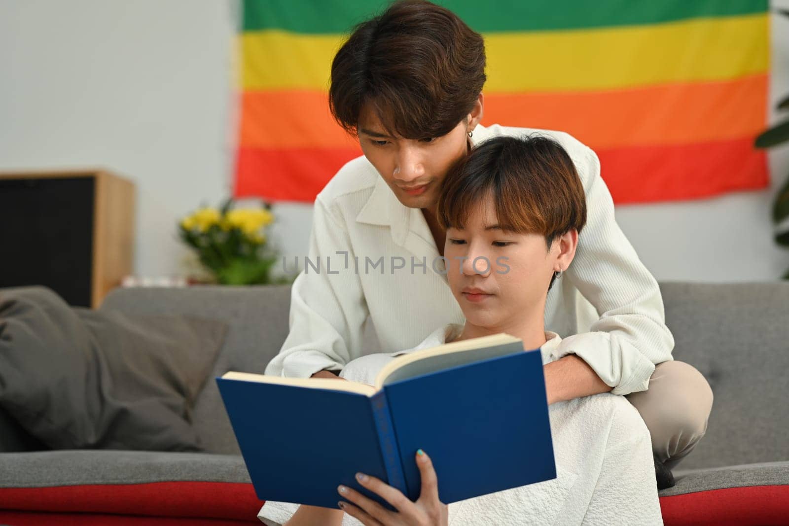 Happy gay couple reading book on sofa at home, spending time together on weekend. LGBTQ people lifestyle and love emotion by prathanchorruangsak