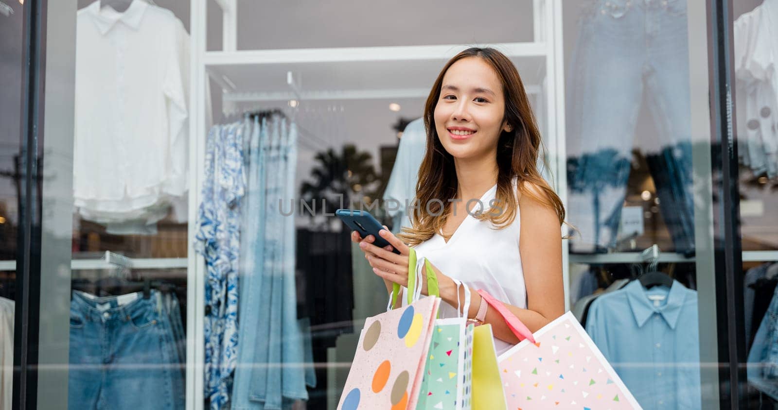 woman holding shopping bags and a cellphone smiles in front of a store window by Sorapop