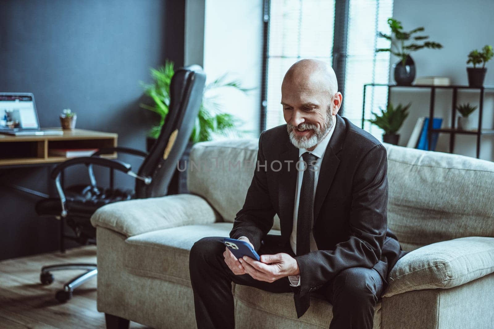 Senior businessman dressed in black suit, sporting warm smile, while browsing internet on phone. Mature man, with distinguished silver beard, seen resting on sofa in loft office. . High quality photo