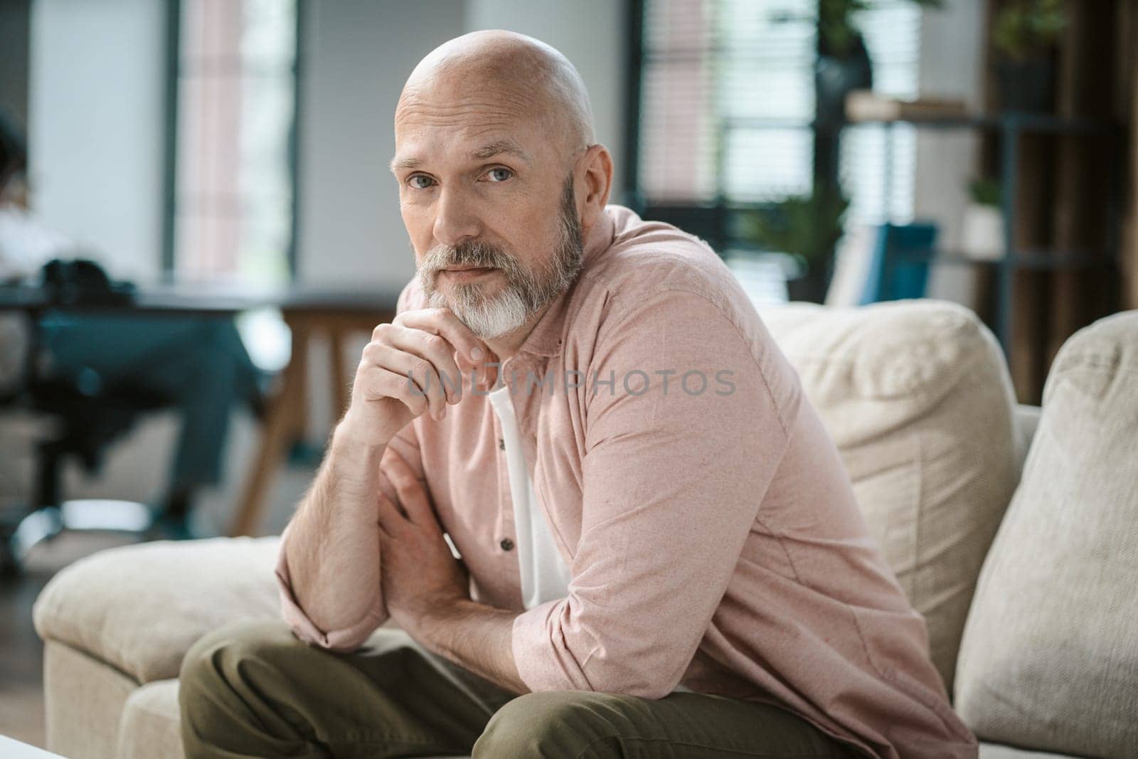 Wise senior man, with touch of grey in beard, looks directly into camera within cozy interior of home. His hand gently rests upon silver beard, exuding sense of thoughtfulness and contemplation. by LipikStockMedia