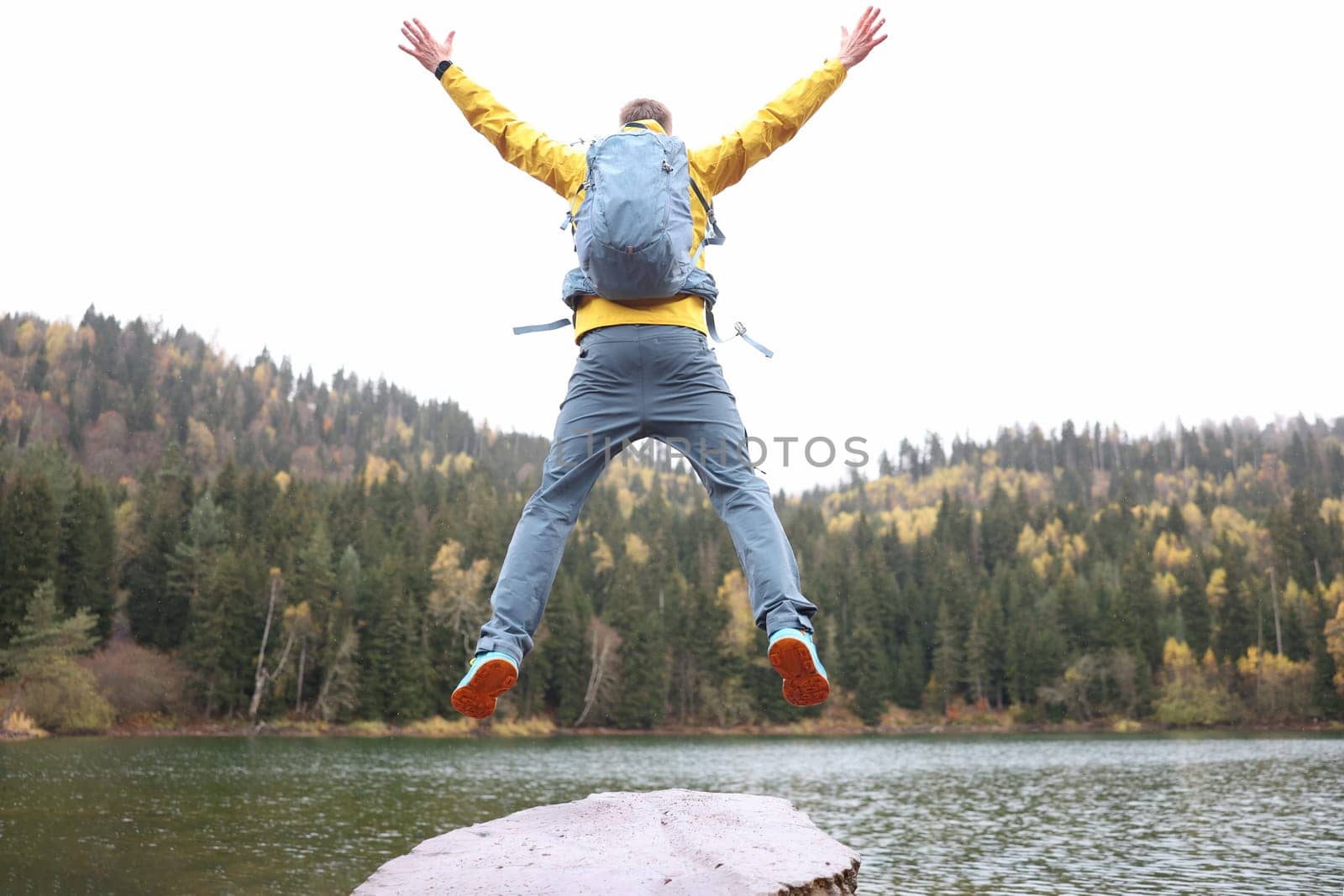 Happy male traveler with backpack jumping near forest lake back view. Travel and goal achievement concept