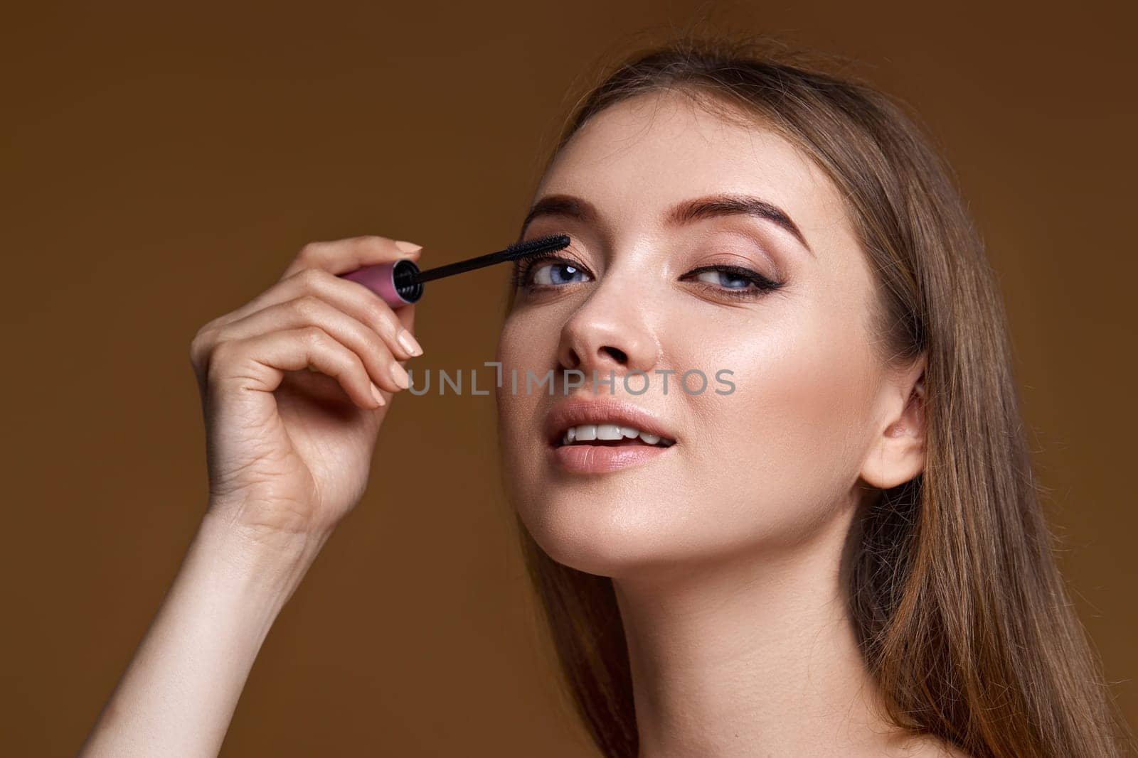 Beautiful woman applying black mascara on long thick eyelashes with brush on beige background