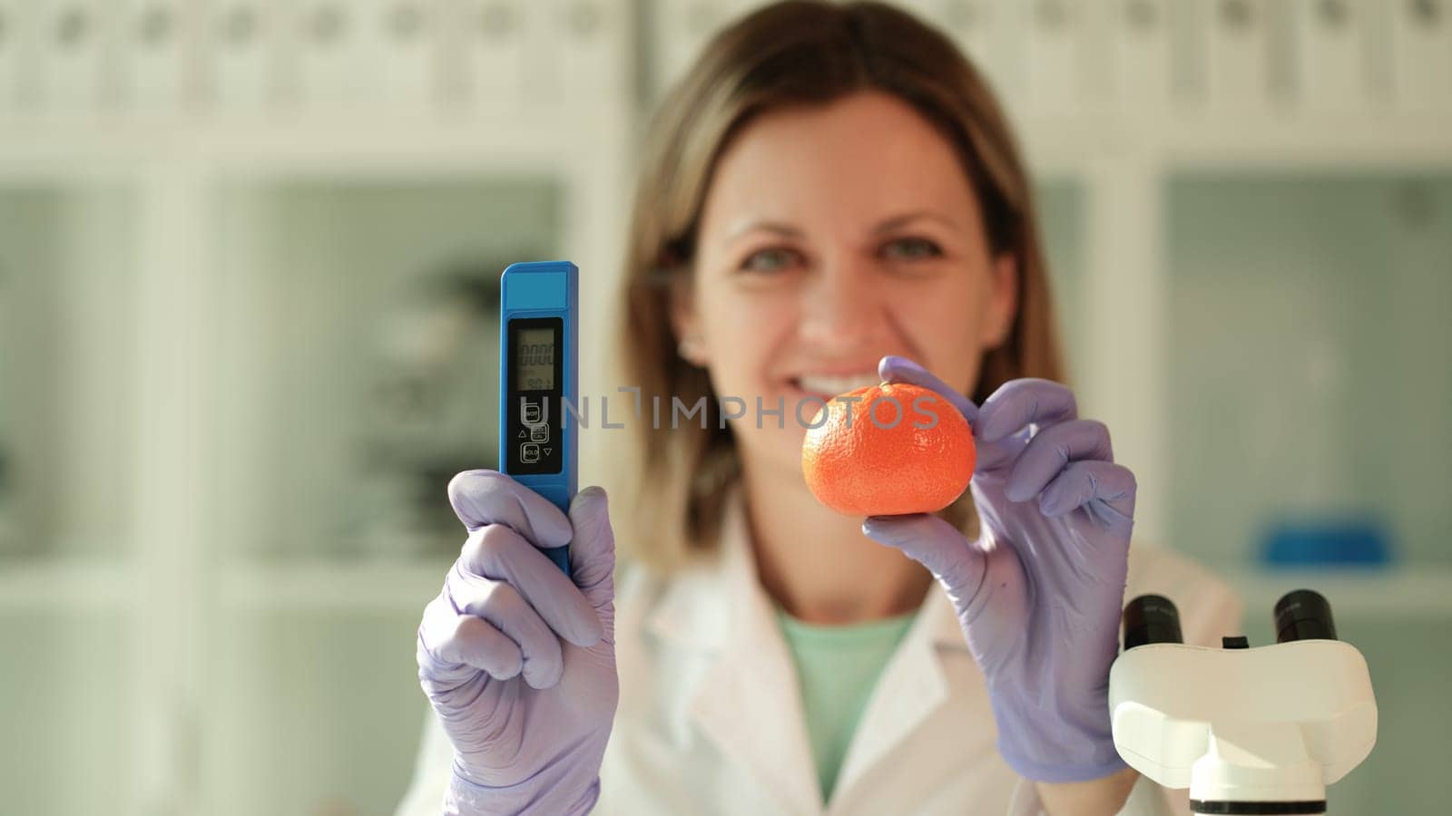 Scientist chemist holding in hands tangerine and device for measuring amount of nitrates in chemical laboratory closeup. Fruit and food quality control concept
