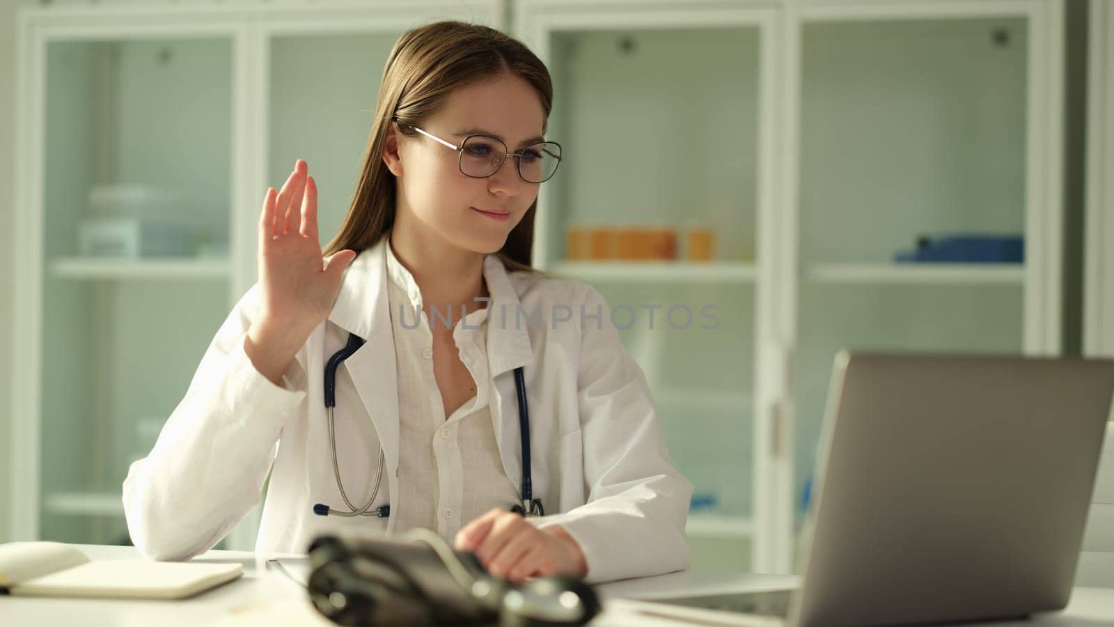 Woman doctor in glasses waving hand at laptop screen in clinic. Telemedicine concept