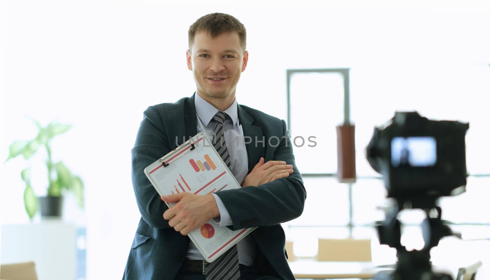 Business coach crossed arms with documents with graphs in front of camera in office. Business development school online concept