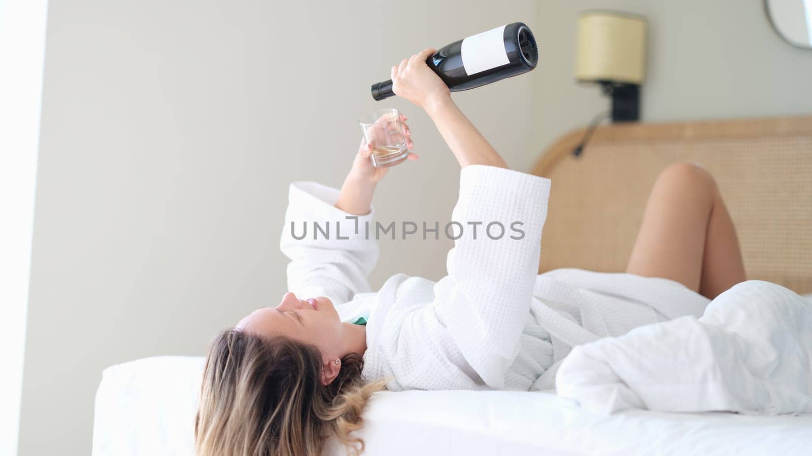 Woman in bathrobe lying in white bed and pouring champagne into glass. Rest and relaxation with alcohol in hotel concept