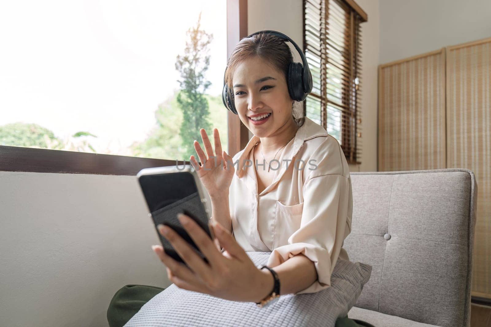 Positive girl in talking on phone in wireless headphones and sitting on couch. Happy woman smiles and has video call with friend.