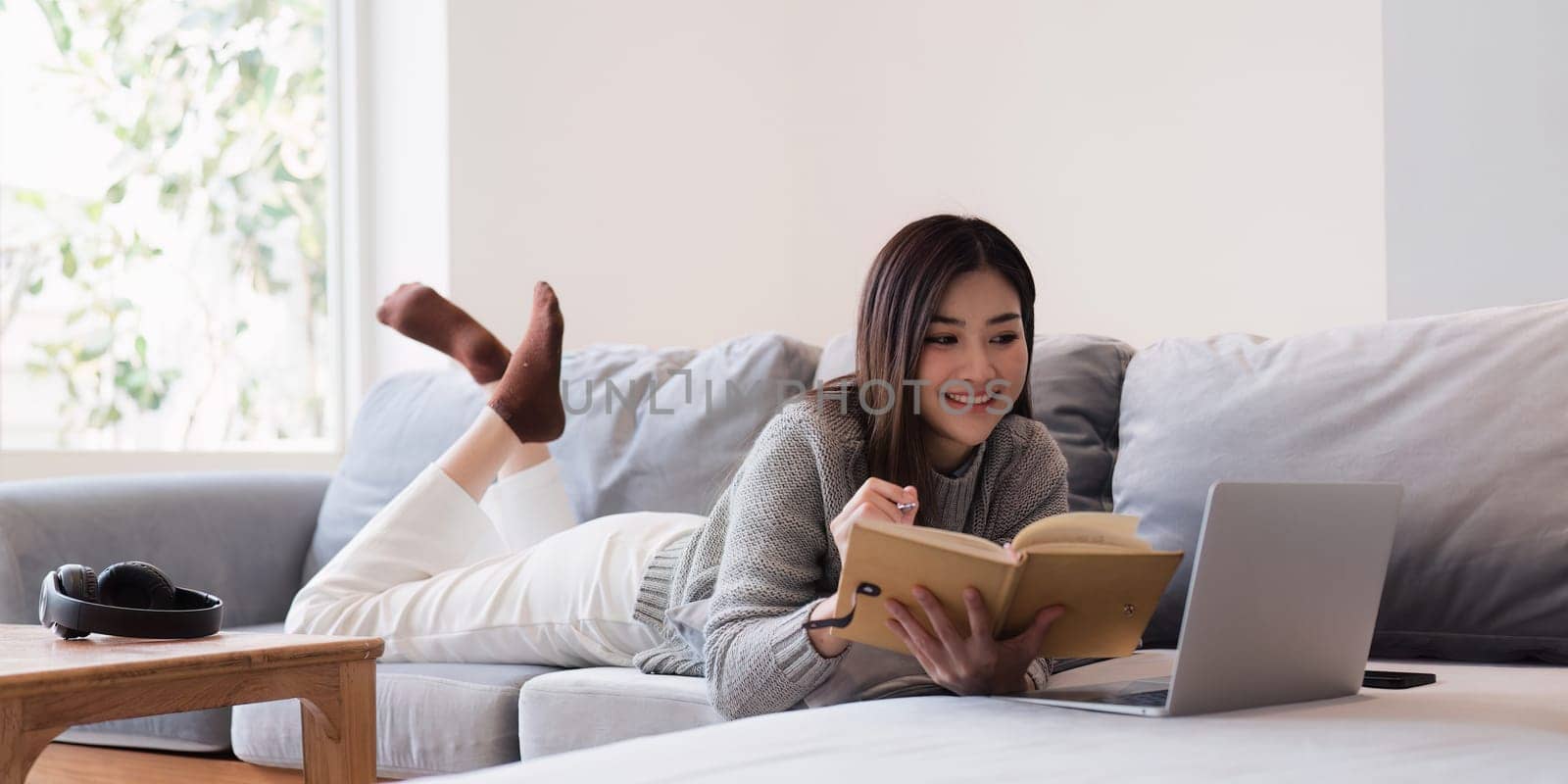 Online education, e-learning. Asian woman in stylish casual clothes, studying using a laptop, listening to online lecture, taking notes, online study at home by itchaznong