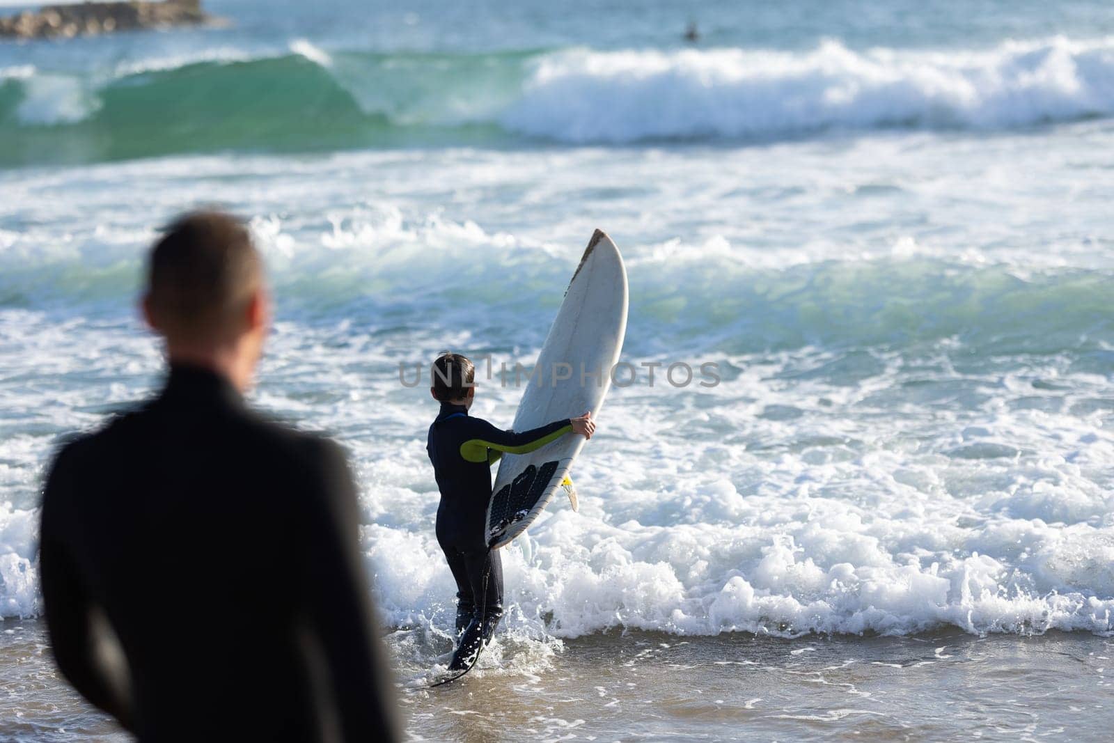 Smiling son on the shoulders of his father walking in the sea wearing wetsuits. Mid shot